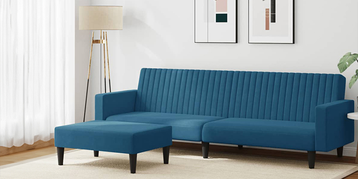 En modern blå sammetssoffa med divan, vertikal kanalsöm och matchande ottoman. Soffan har svarta träben och står i ett ljust vardagsrum med inramad väggkonst och en golvlampa i hörnet.
